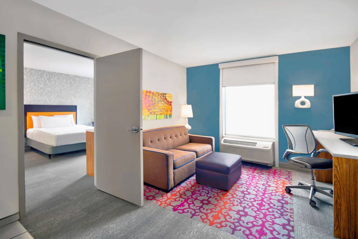 A hotel room with a bed, a sofa, an ottoman, a desk with a chair, a TV, and a window with blinds.
