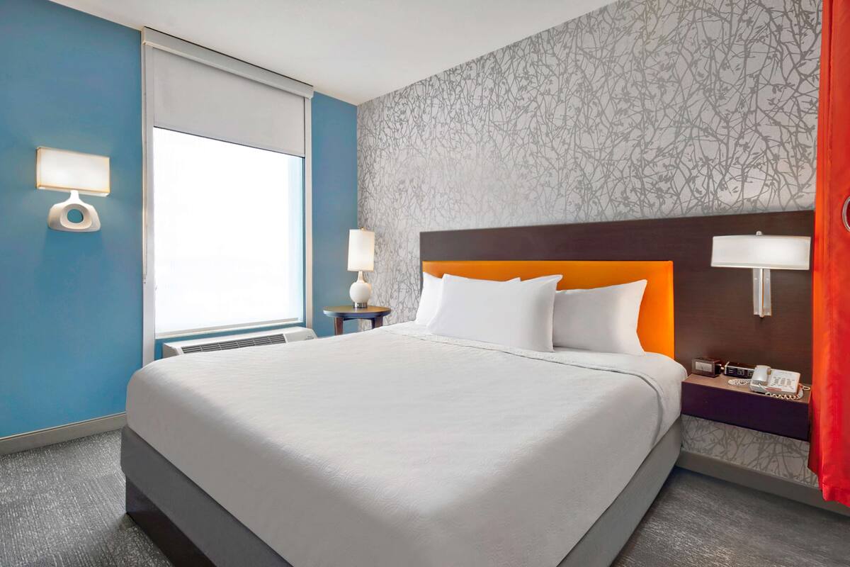 A hotel room with a large bed, two bedside tables with lamps, a window with blinds, and a patterned wall.