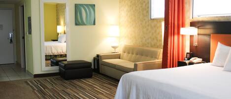 A hotel room with a bed, a sofa, a chair, a small table, a nightstand, a lamp, and a mirror.