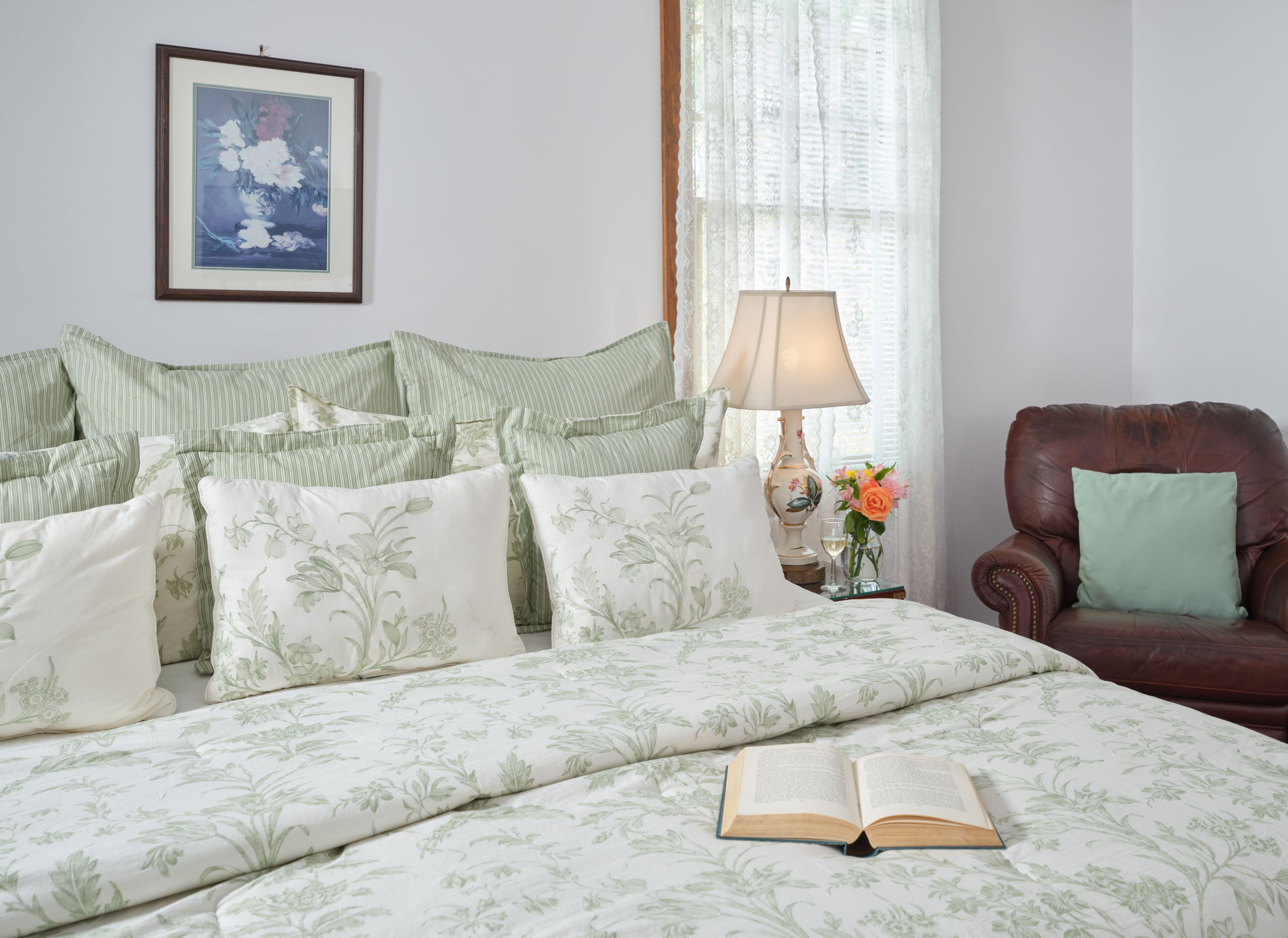 Una cama bien hecha con ropa de cama floral, un sillón de cuero marrón, una mesita de noche con lámpara y flores, y un cuadro en la pared.