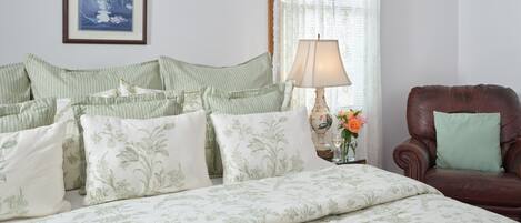 A neatly made bed with floral bedding, a brown leather armchair, a bedside table with a lamp and flowers, and a framed picture on the wall.