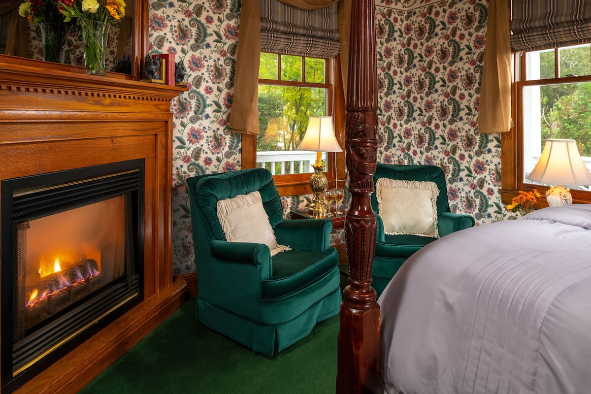 A room with a fireplace, two green armchairs, a four-poster bed, and floral wallpaper.