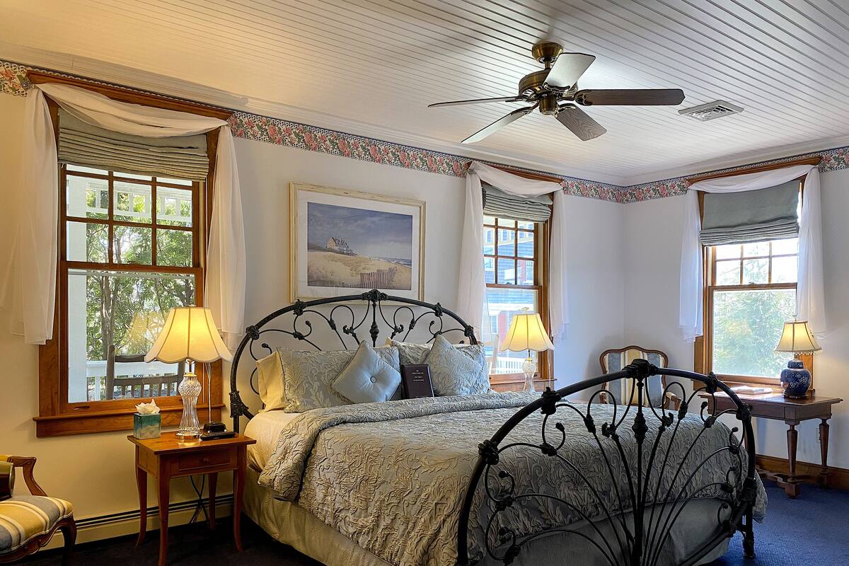 A bedroom with a four-poster bed, two bedside tables with lamps, a ceiling fan, and large windows with curtains.
