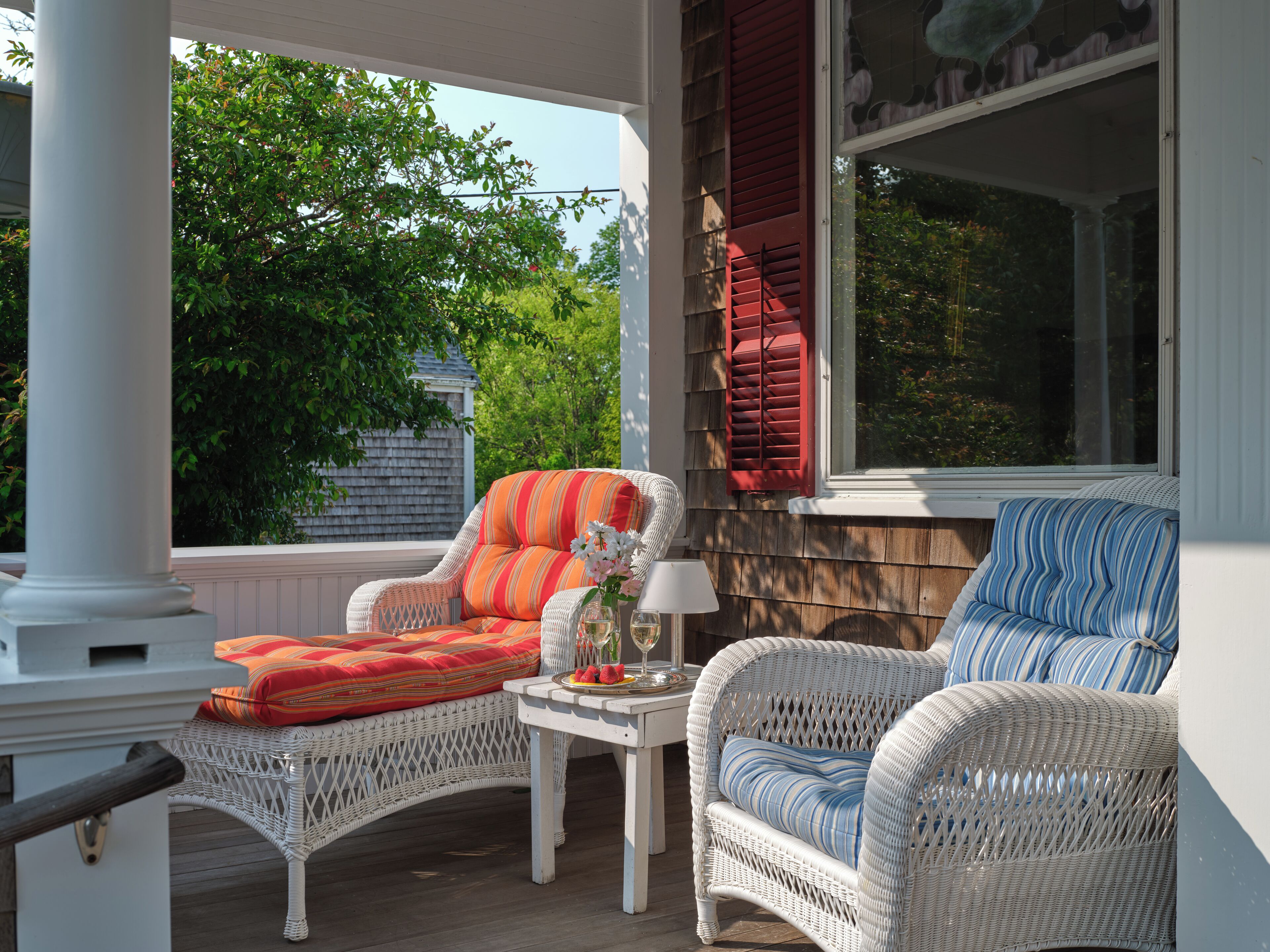 A covered porch with wicker furniture, including a chair and a sofa, and a small table with a vase and a glass.