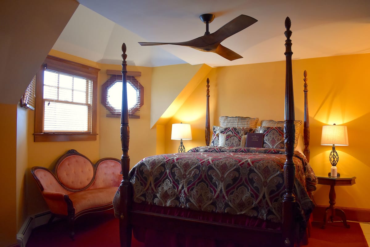 A four-poster bed with a ceiling fan, a chaise lounge, and two bedside lamps in a room with yellow walls and a window with blinds.