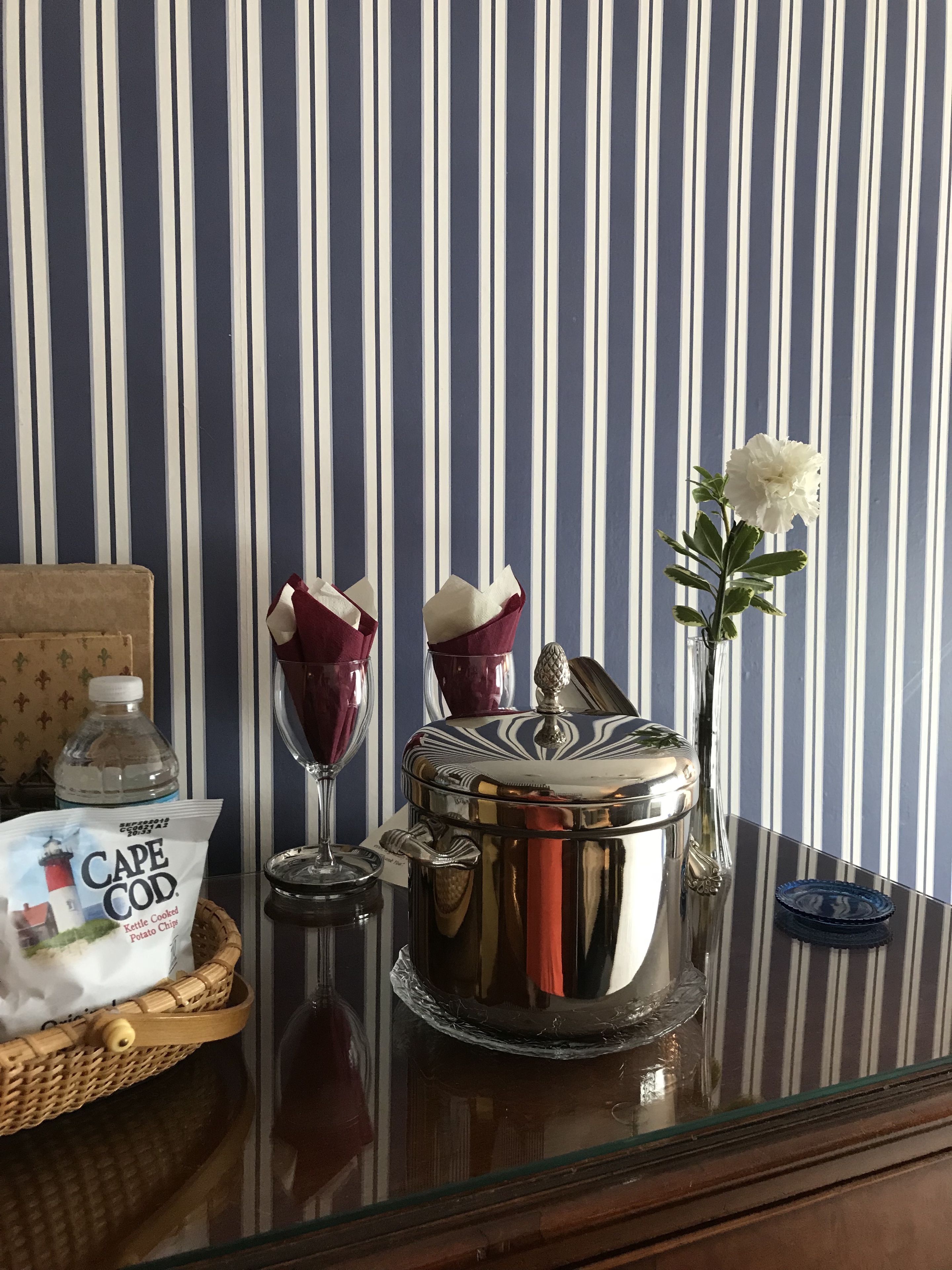 A table with a bottle of water, a basket of Cape Cod potato chips, a glass with a napkin, a silver pot, and a vase with a white flower.