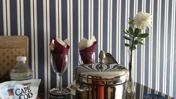 A table with a bottle of water, a basket of Cape Cod potato chips, a glass with a napkin, a silver pot, and a vase with a white flower.
