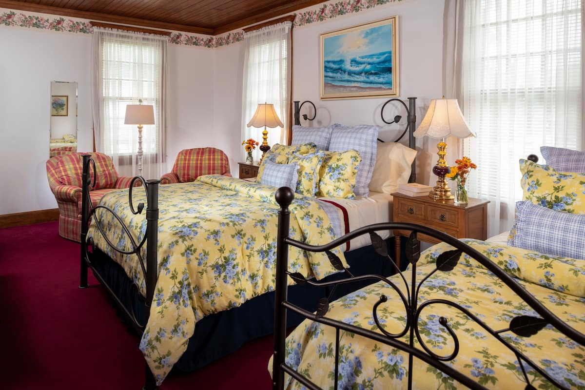 A bedroom with two beds, floral bedding, a wooden headboard, a nightstand with lamps, and a painting on the wall.