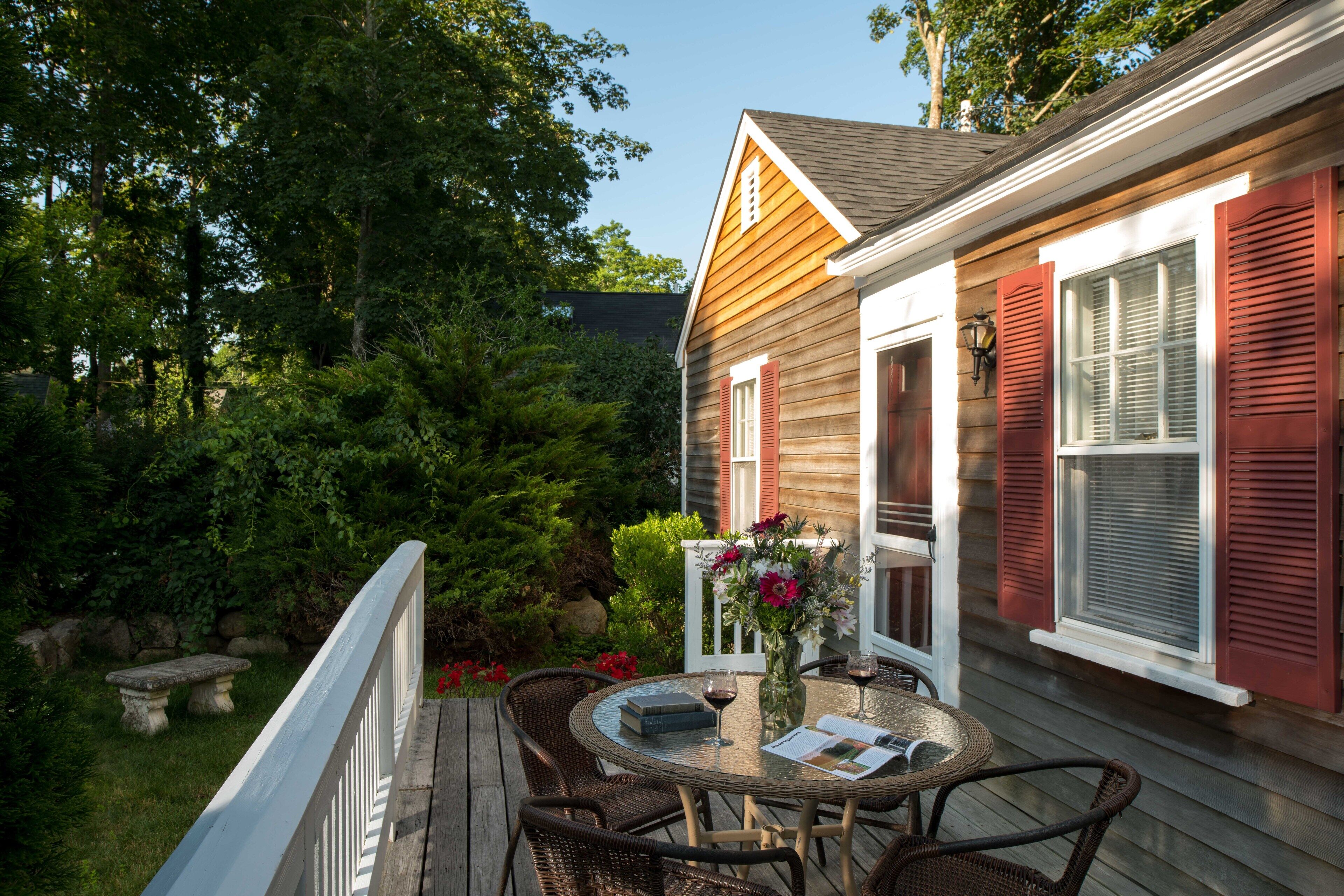 A wooden deck with wicker chairs and a table, a white railing, and a house with red shutters.