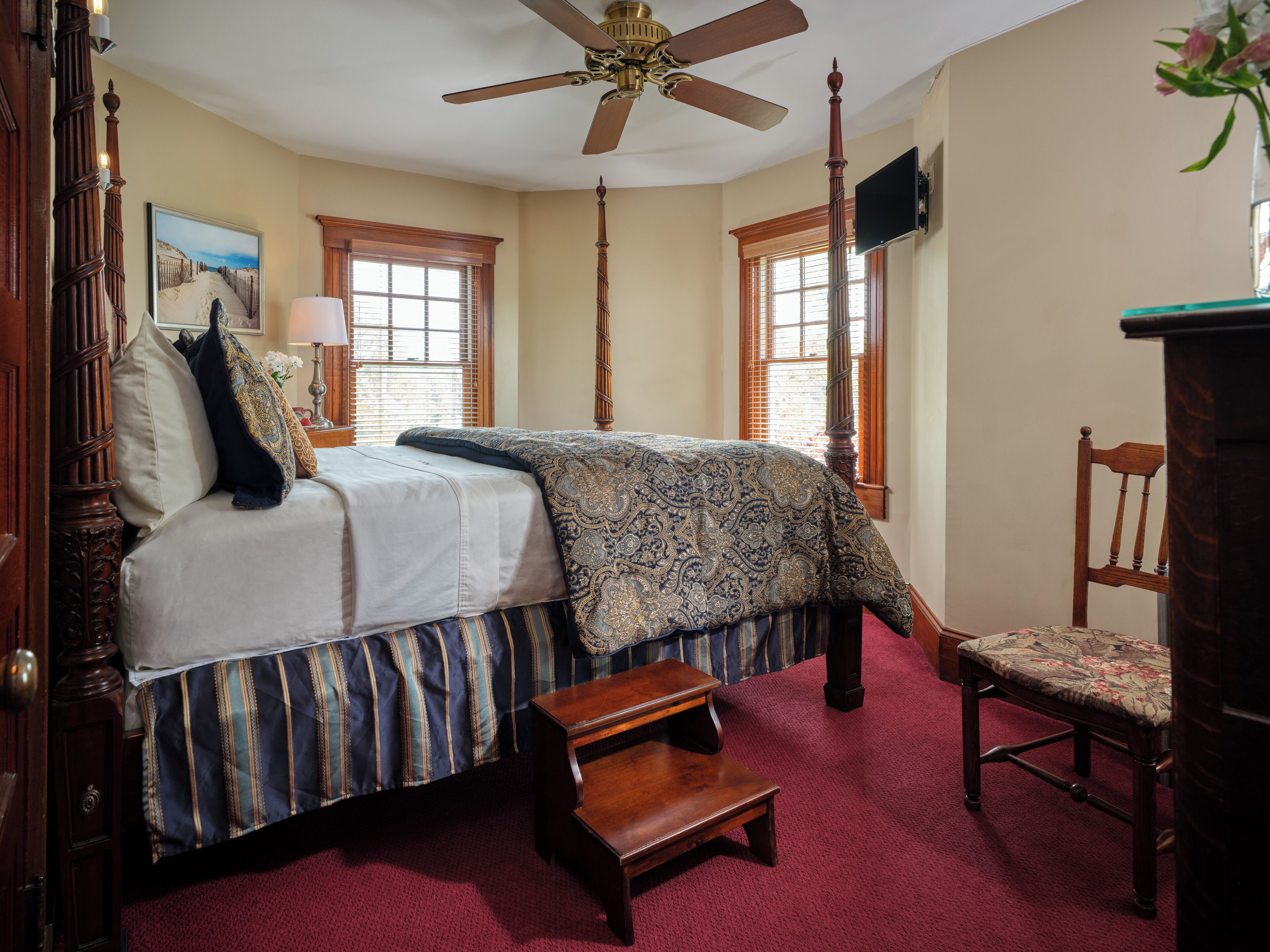 A bedroom with a bed, a wooden chair, a small table, a ceiling fan, and a TV mounted on the wall.