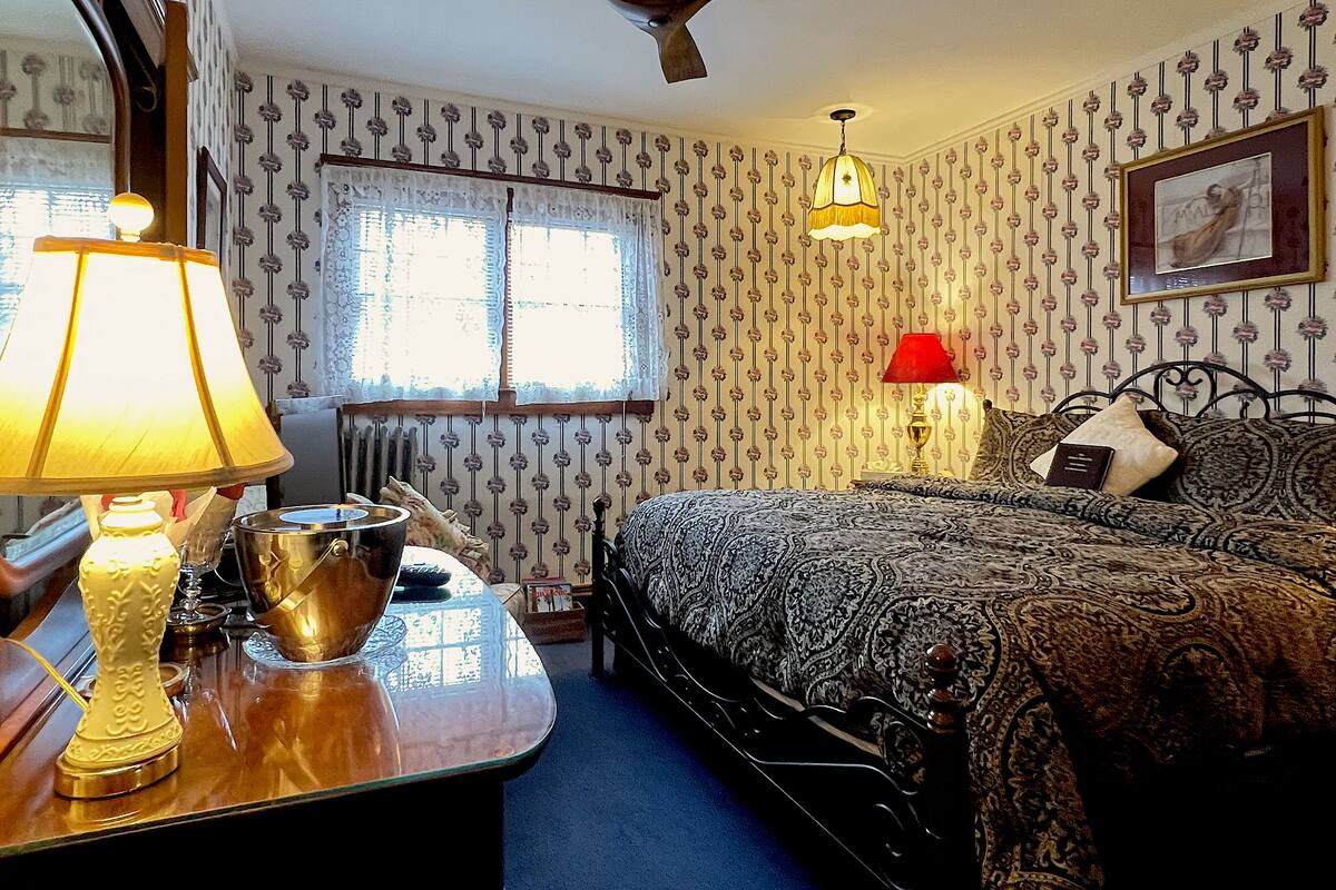 A bedroom with a bed, two bedside lamps, a ceiling fan, a window with lace curtains, and patterned wallpaper.