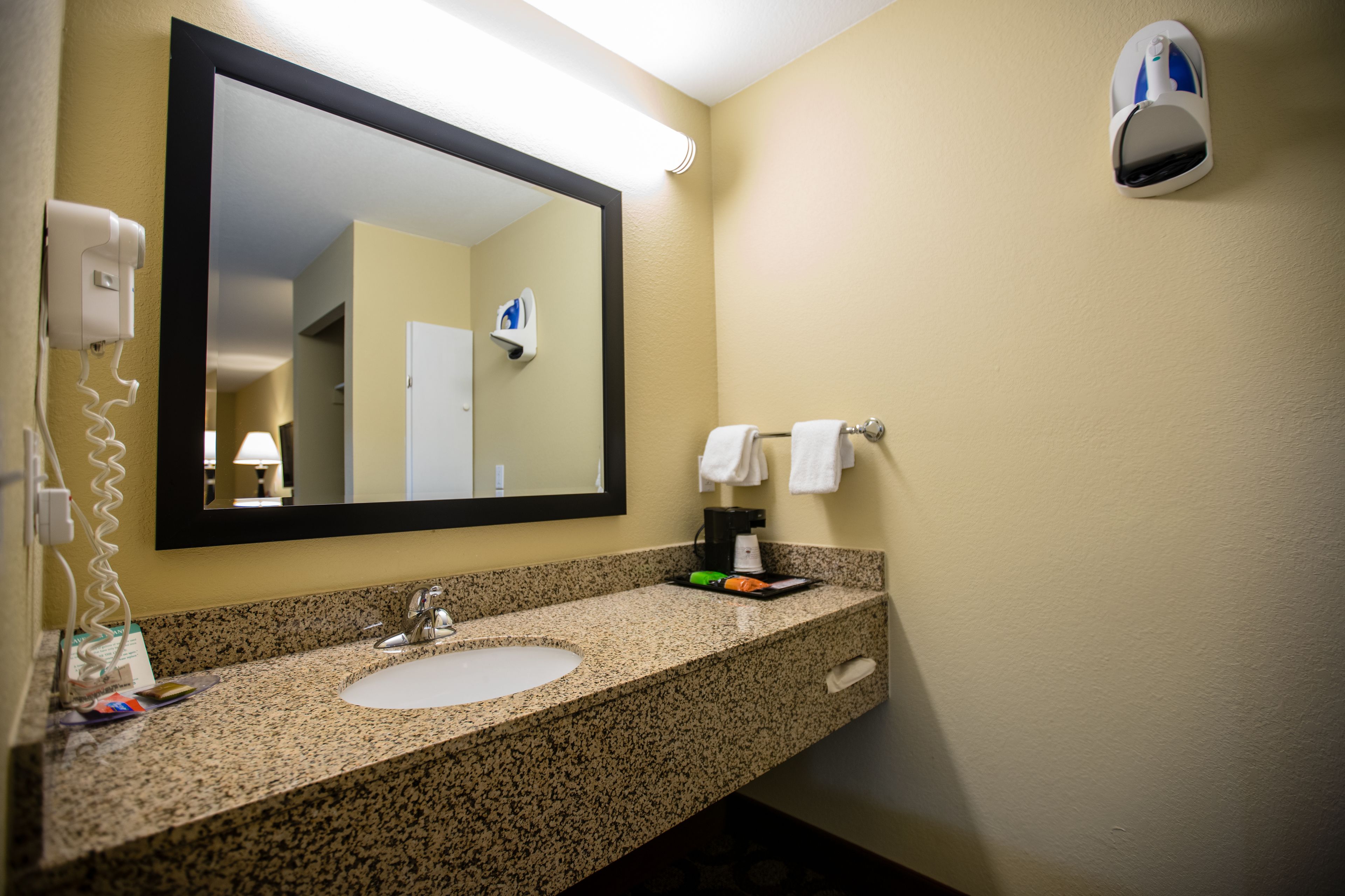 A hotel bathroom with a granite countertop sink, a large mirror, a hair dryer, and a towel rack.