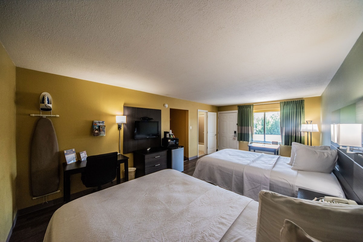 Quarto de hotel com duas camas, uma mesa de trabalho com cadeira, televisão e uma prancha de surf na parede.