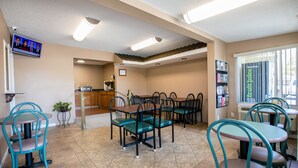 A dining area with multiple tables and chairs, a kitchen in the background, and a television mounted on the wall.