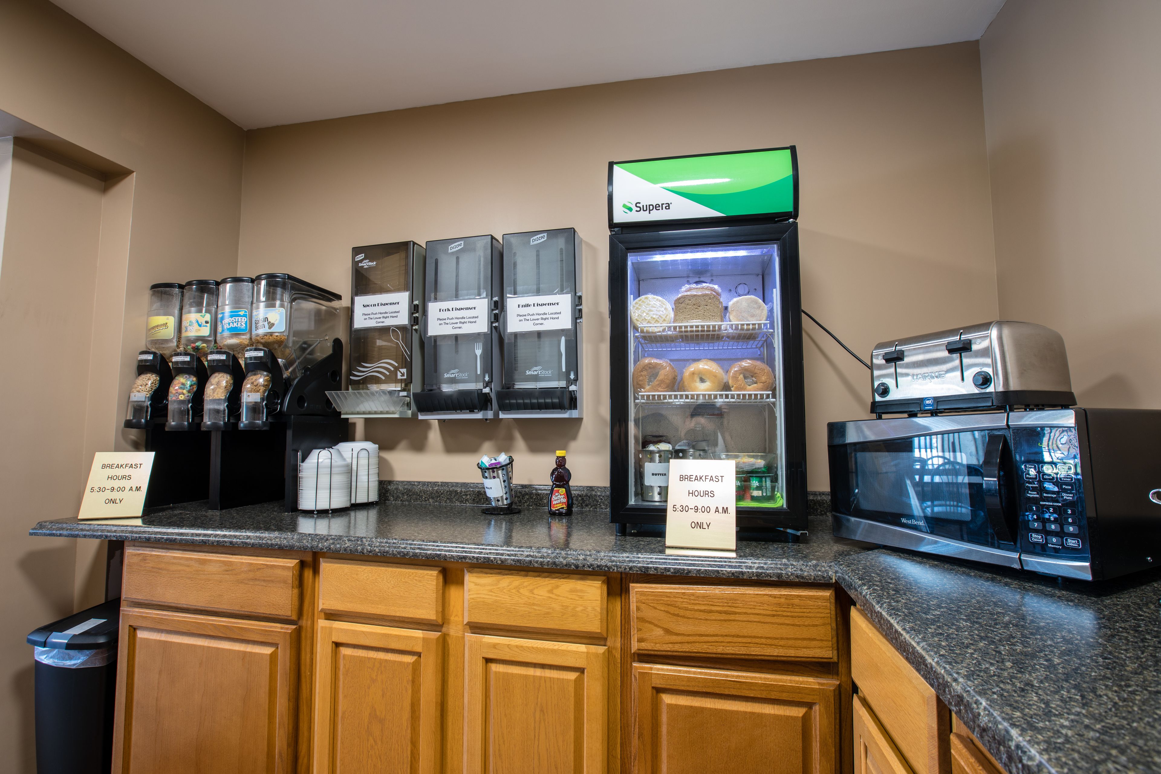 A breakfast bar with a coffee machine, a refrigerator with pastries, a toaster, and a sign indicating breakfast hours.