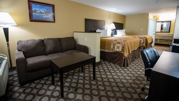 A hotel room with a sofa, a small table, a bed with a patterned bedspread, a desk, a chair, a lamp, a heater, a mirror, and a framed picture on the wall.
