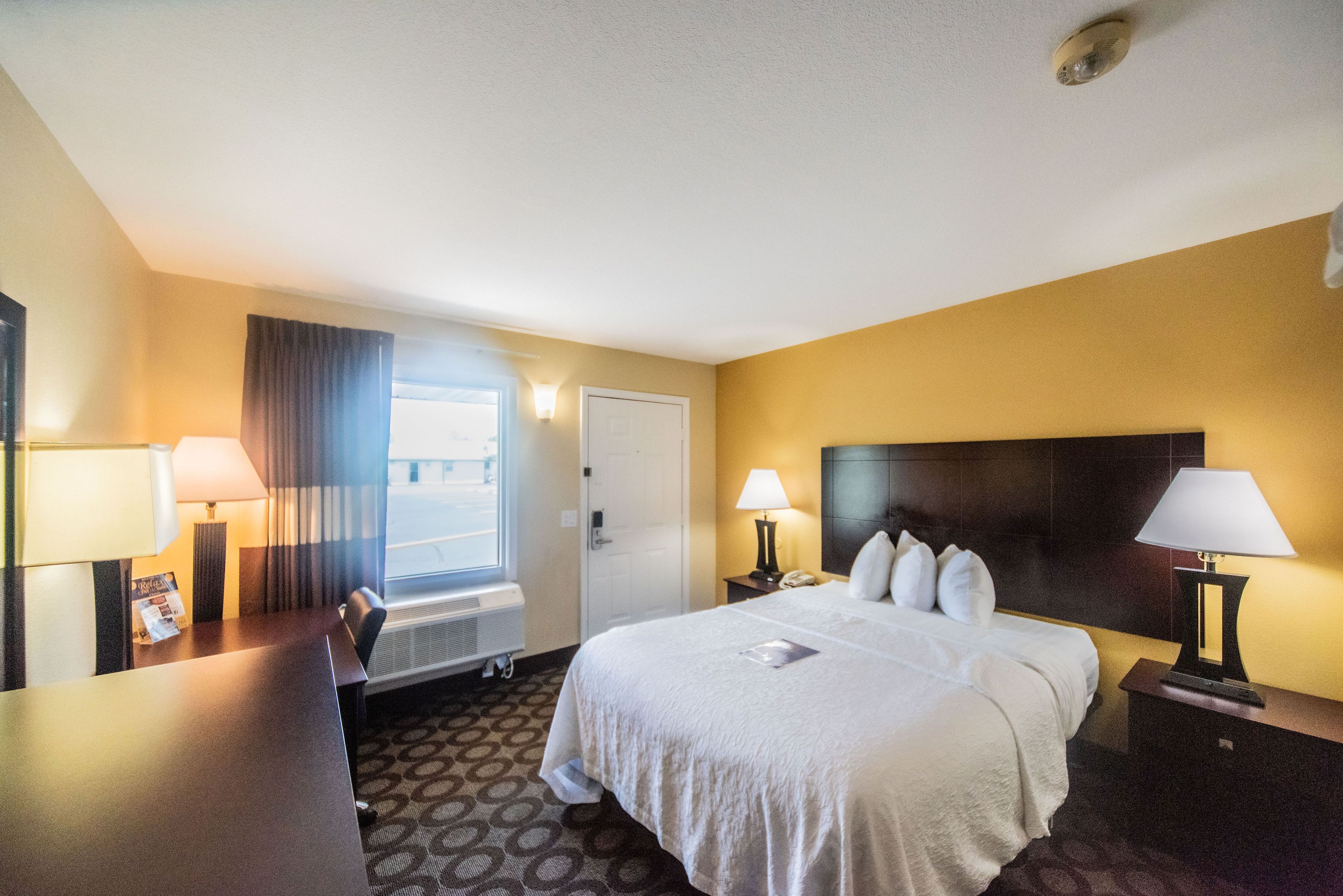 A hotel room with a large bed, two bedside tables with lamps, a desk, a chair, and a window with curtains.