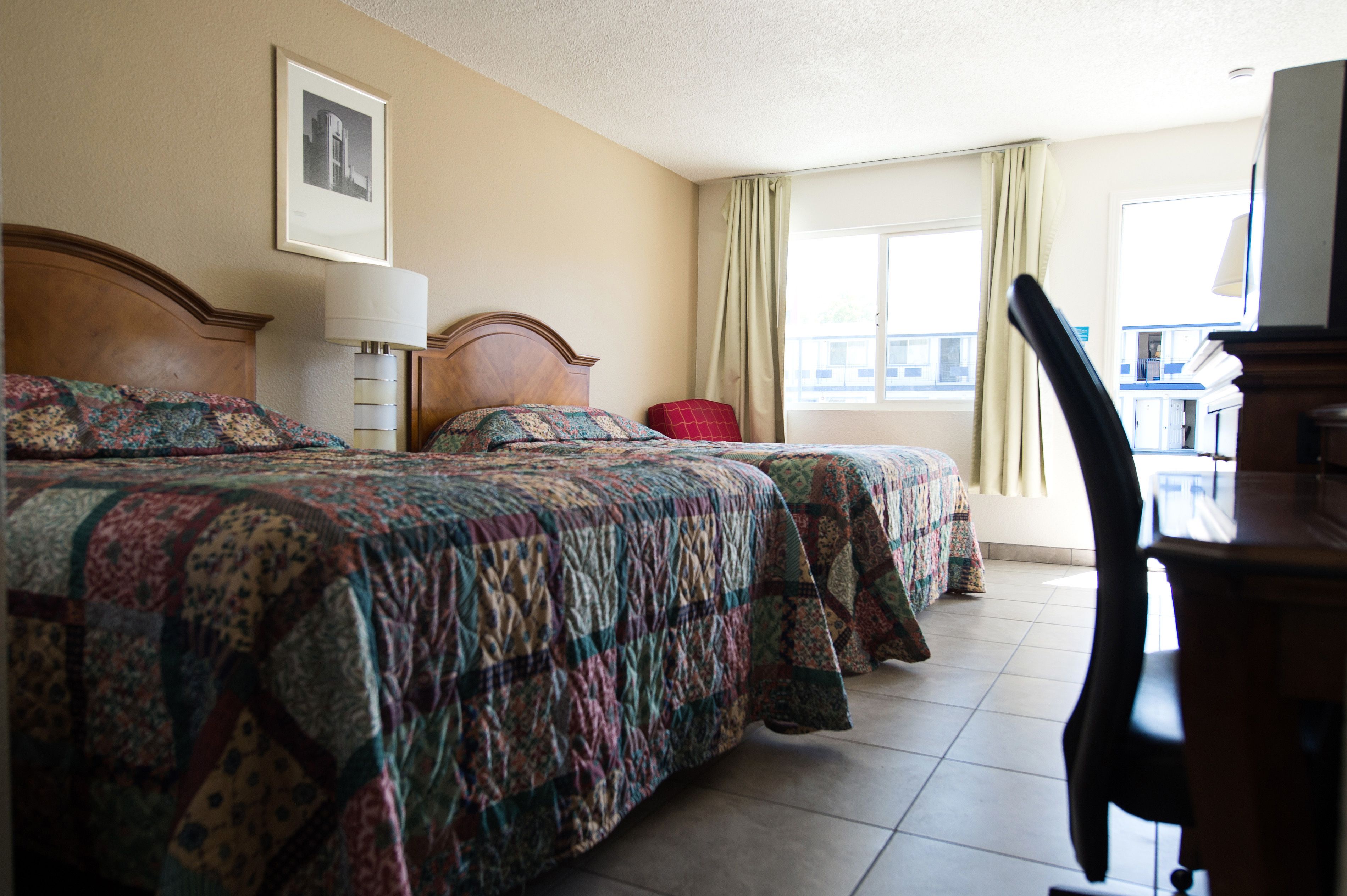 Quarto de hotel com duas camas, uma escrivaninha, uma cadeira, janela com cortinas e um quadro na parede.