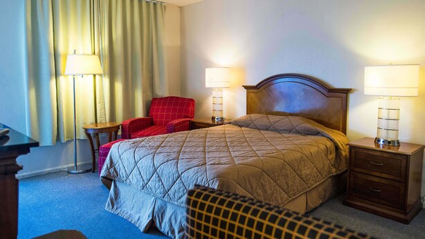 Una habitación de hotel con una cama, dos mesitas de noche con lámparas, un sillón rojo y un escritorio con lámpara.