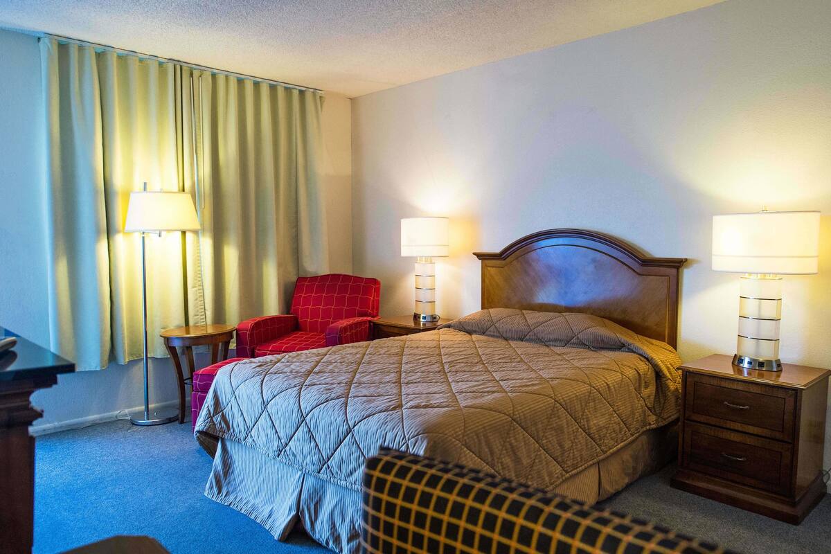 A hotel room with a bed, two bedside tables with lamps, a red armchair, and a desk with a lamp.