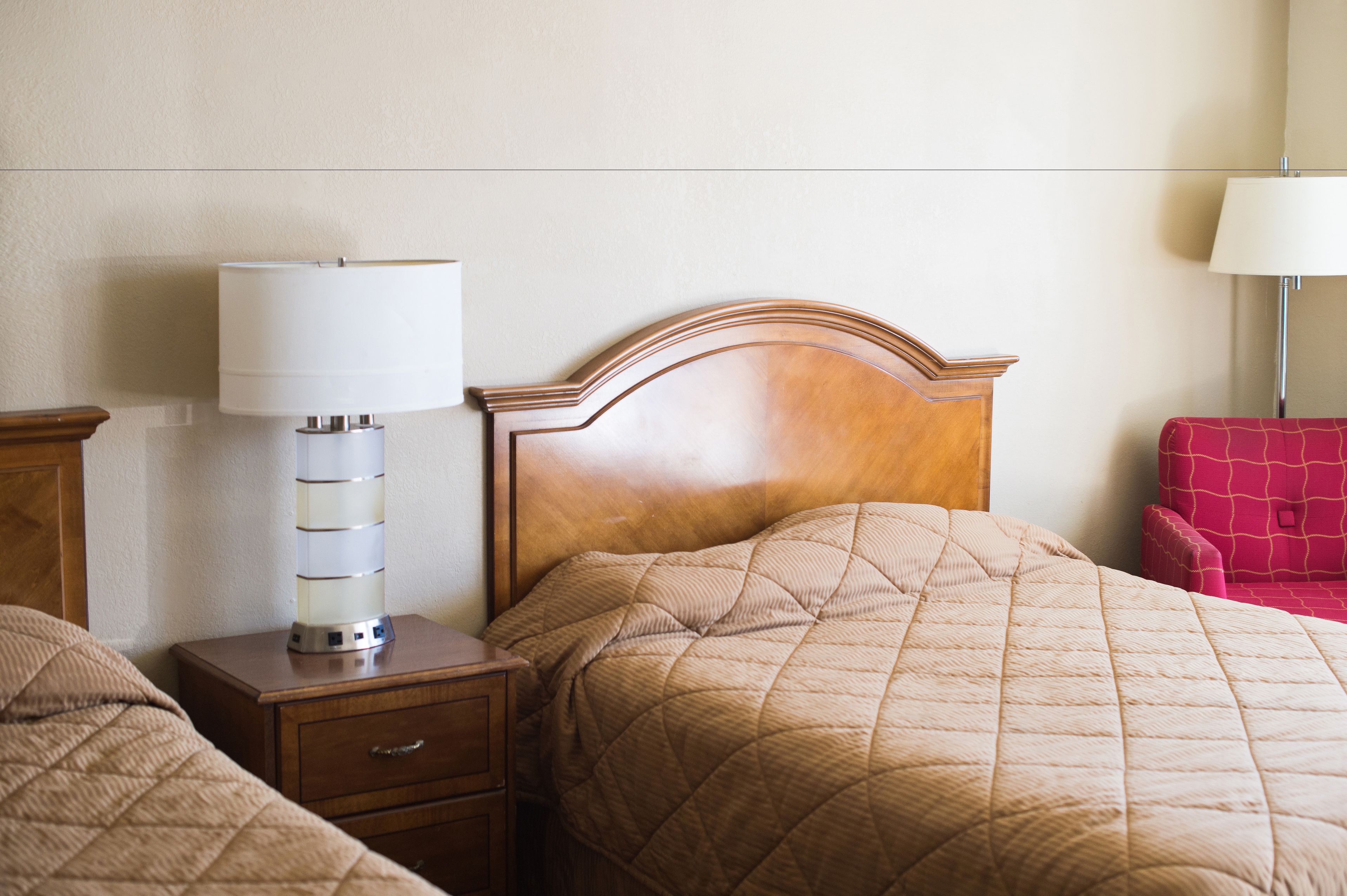 Une chambre d’hôtel avec une tête de lit en bois, une table de chevet avec une lampe, une chaise rouge et un lit recouvert d’une courtepointe beige.