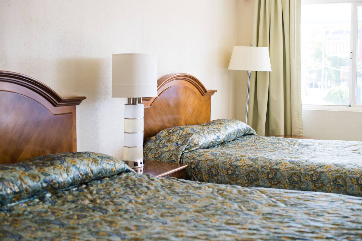 A hotel room with two beds, a nightstand, a lamp, and a window with curtains.