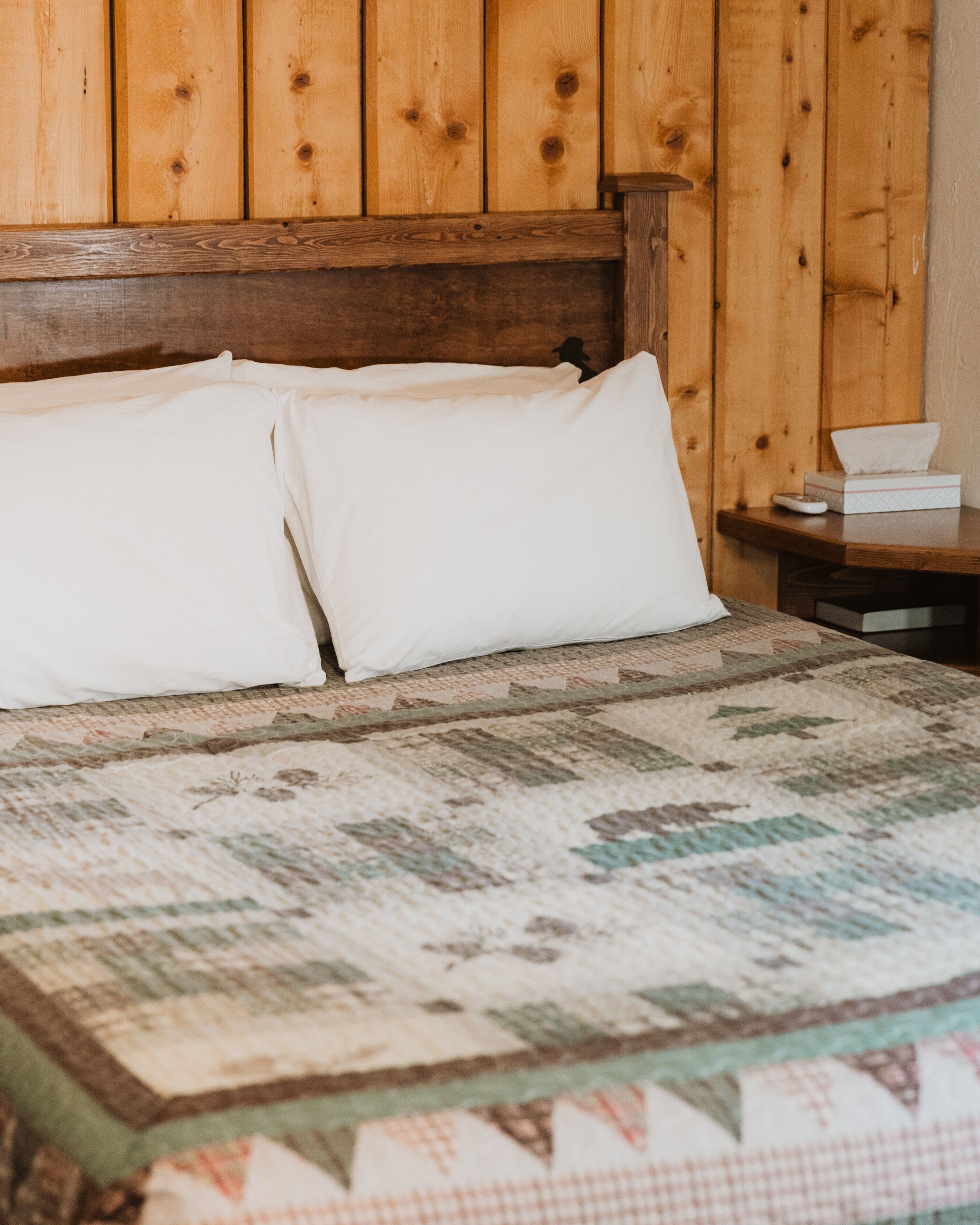 A bed with two white pillows and a quilt, a wooden headboard, and a nightstand with a tissue box.