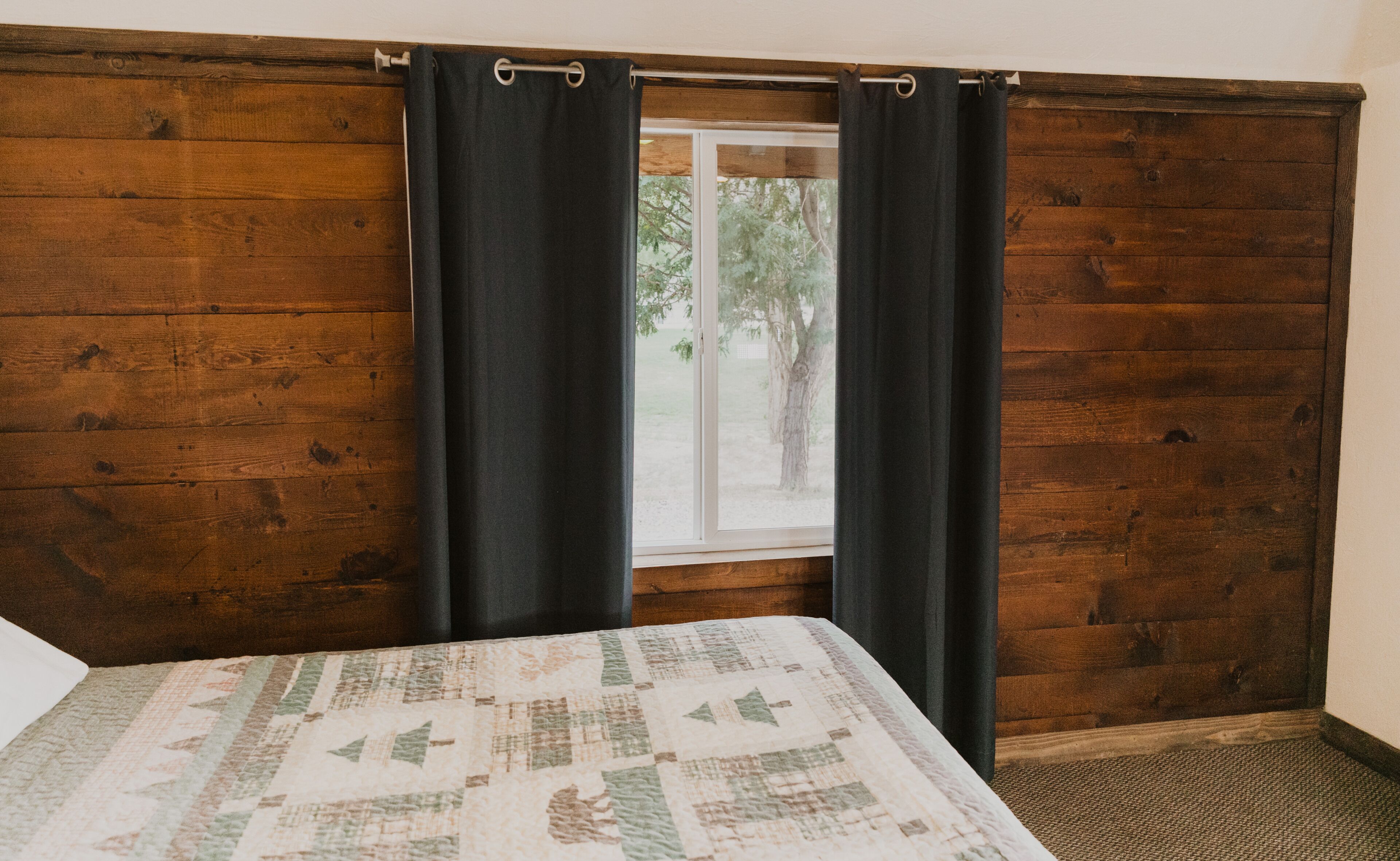 A room with a wooden wall, a window with black curtains, and a bed with a quilt.