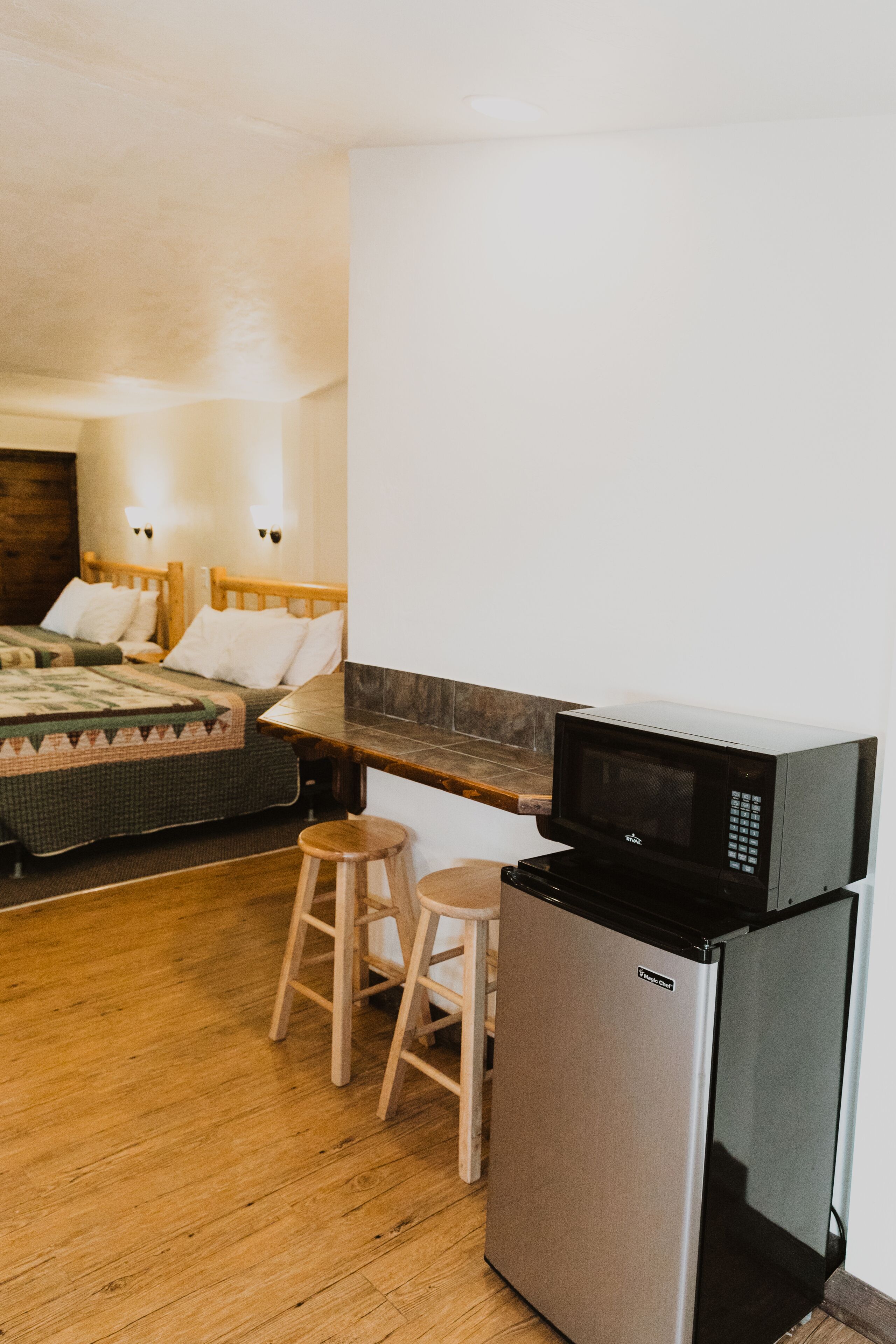 A compact room with a kitchenette, including a microwave and refrigerator, a wooden bed with a quilt, and a small stool.