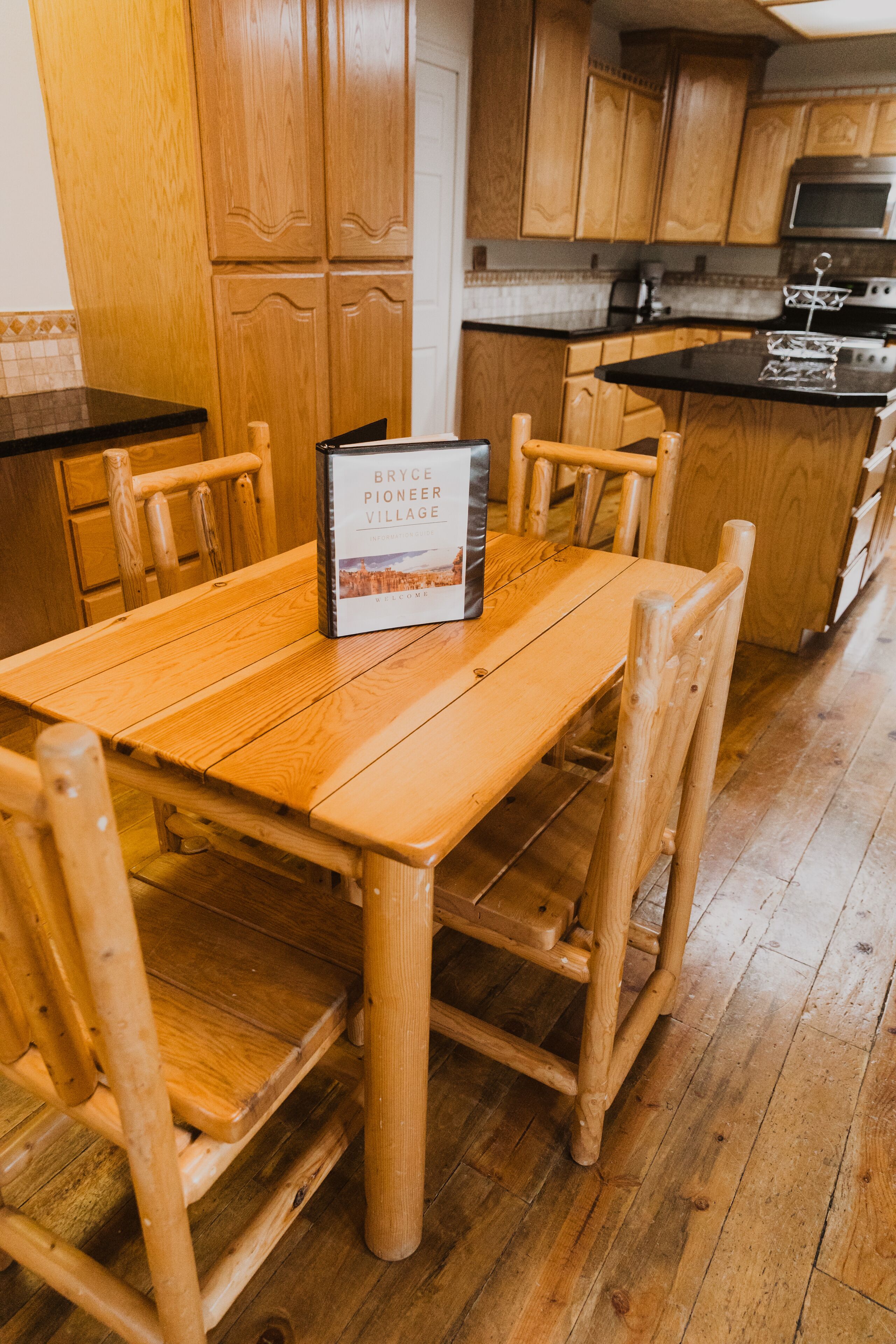 Una mesa de comedor de madera con sillas, un libro titulado "Bryce Pioneer Village" y armarios de madera en una cocina.