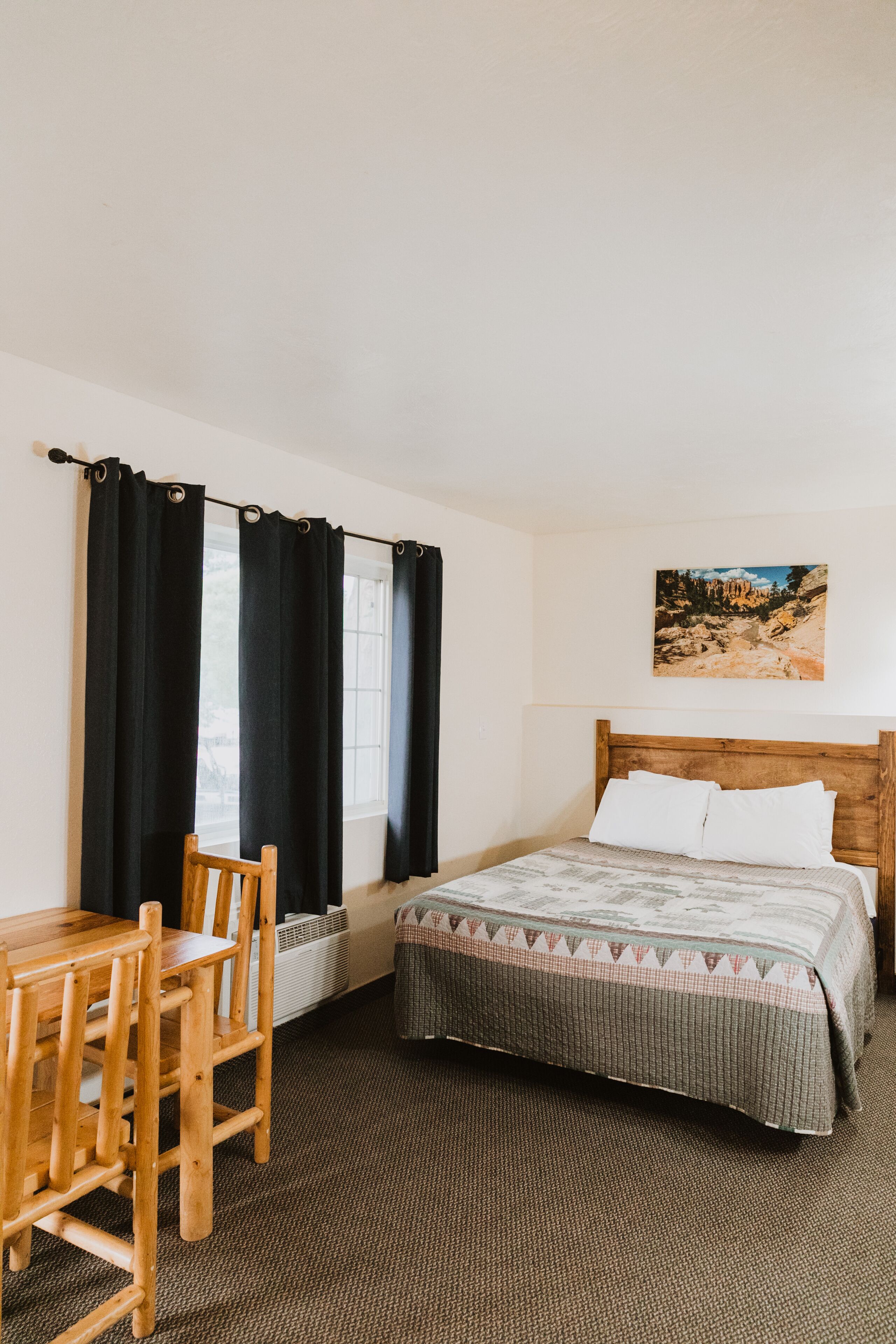 A bedroom with a wooden bed, a desk with a chair, black curtains, a window, and a painting on the wall.
