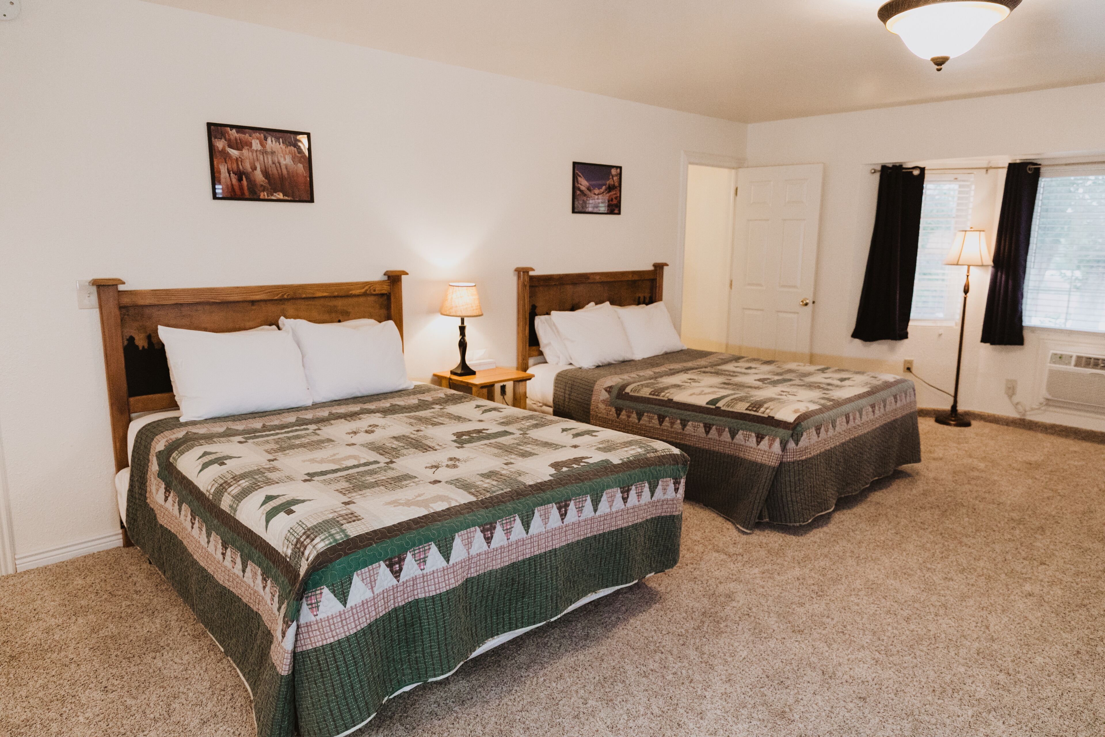 A room with two beds, each with a quilted bedspread, a nightstand with a lamp, a door, and a window with curtains.