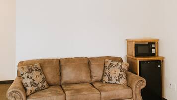 A brown leather sofa with floral cushions in a room with a carpeted floor and a small wooden cabinet with a microwave.