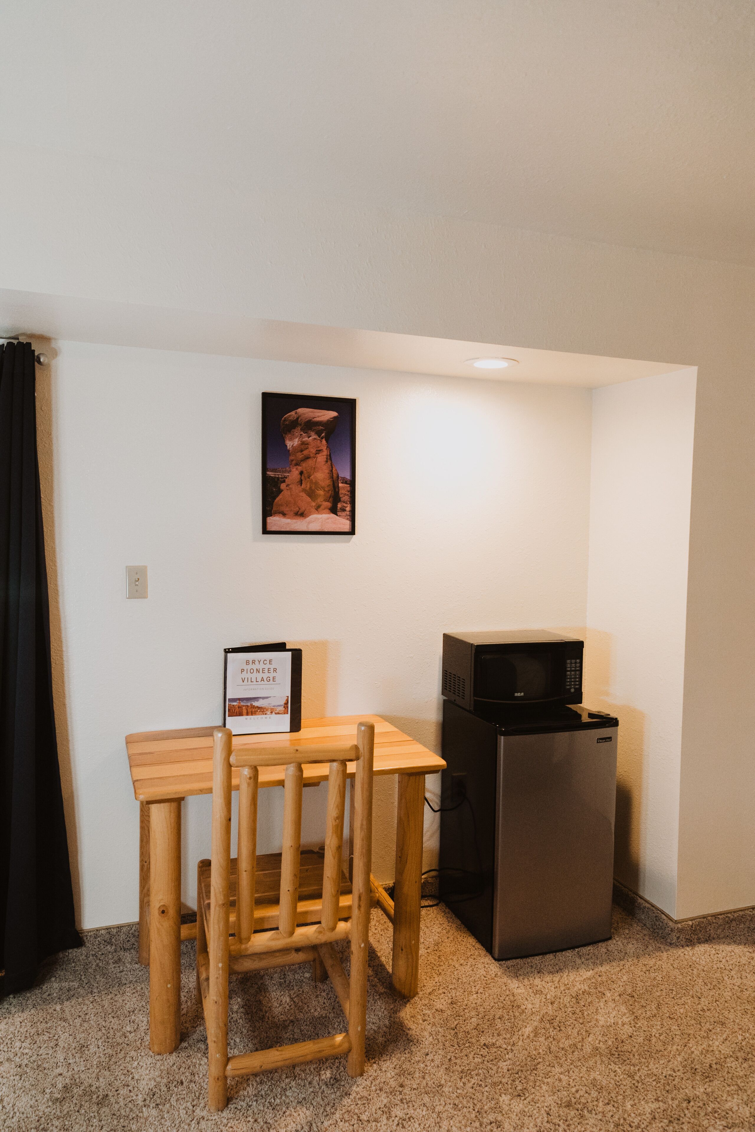 A small kitchenette with a microwave, a refrigerator, a wooden table, and a chair. There is a framed picture on the wall.