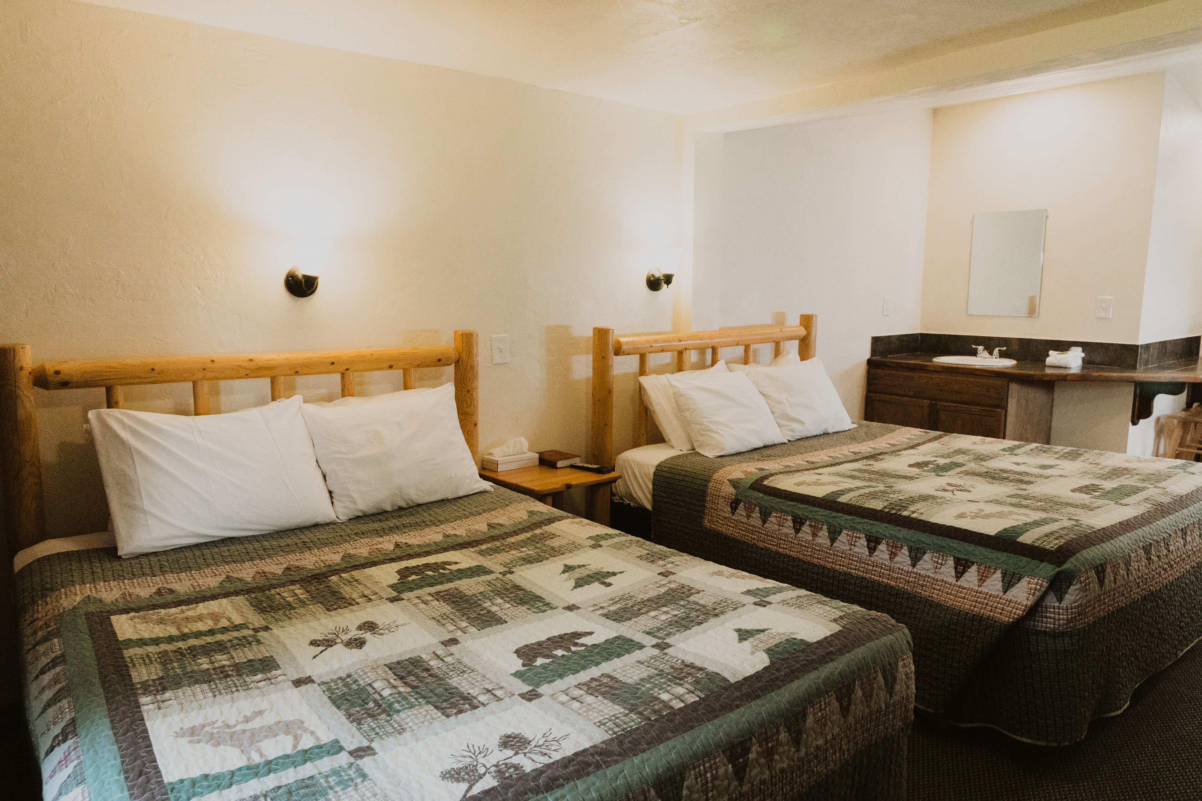 A hotel room with two beds, each with a wooden headboard and white bedding. There is a nightstand with a lamp and a small table between the beds. A bathroom with a sink and mirror is visible in the background.