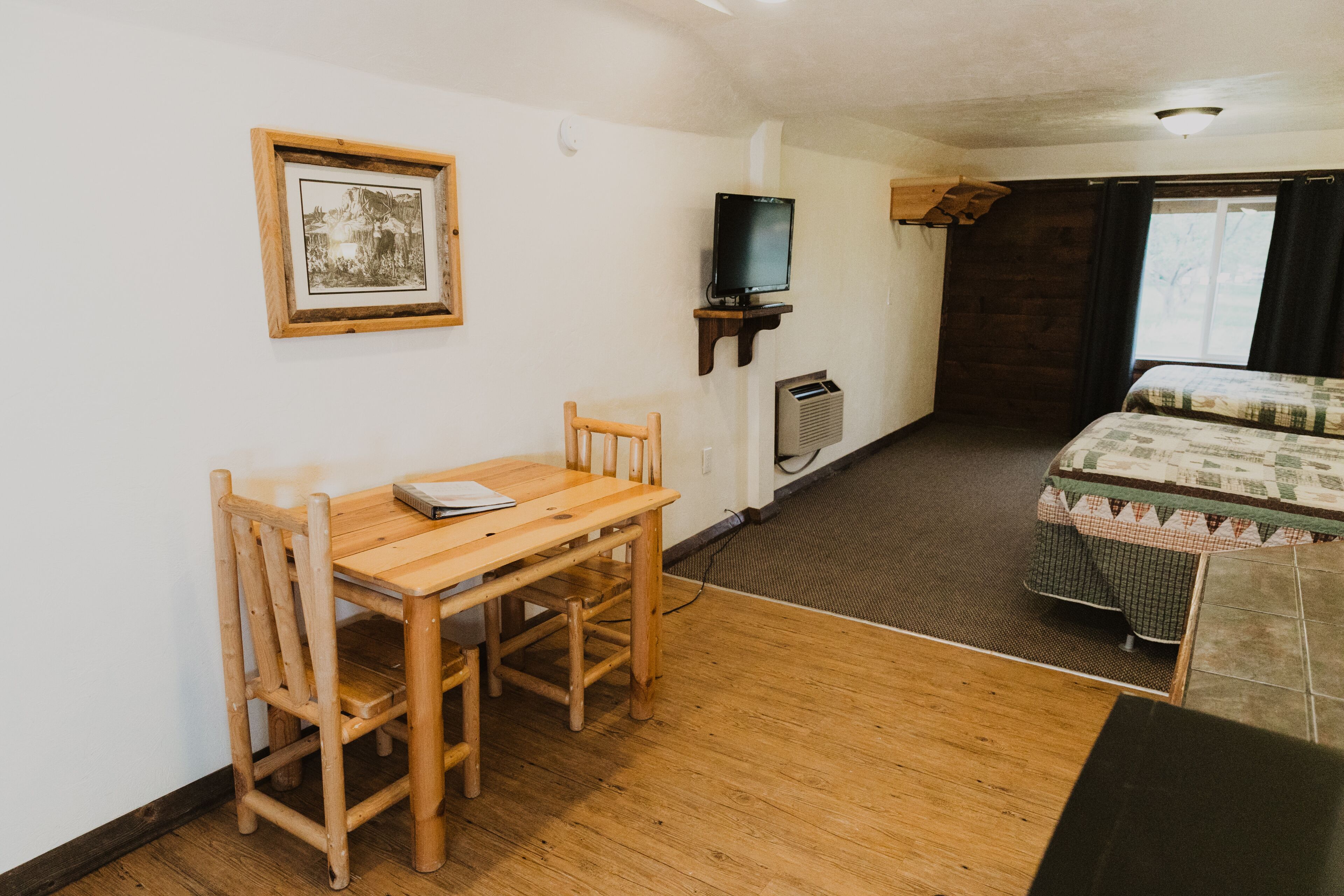 A room with a wooden table and chairs, a television on a stand, a bed with a quilt, and a framed picture on the wall.