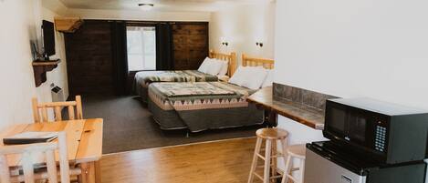 A compact room with a bed, a small kitchenette, a wooden table, and a stool.