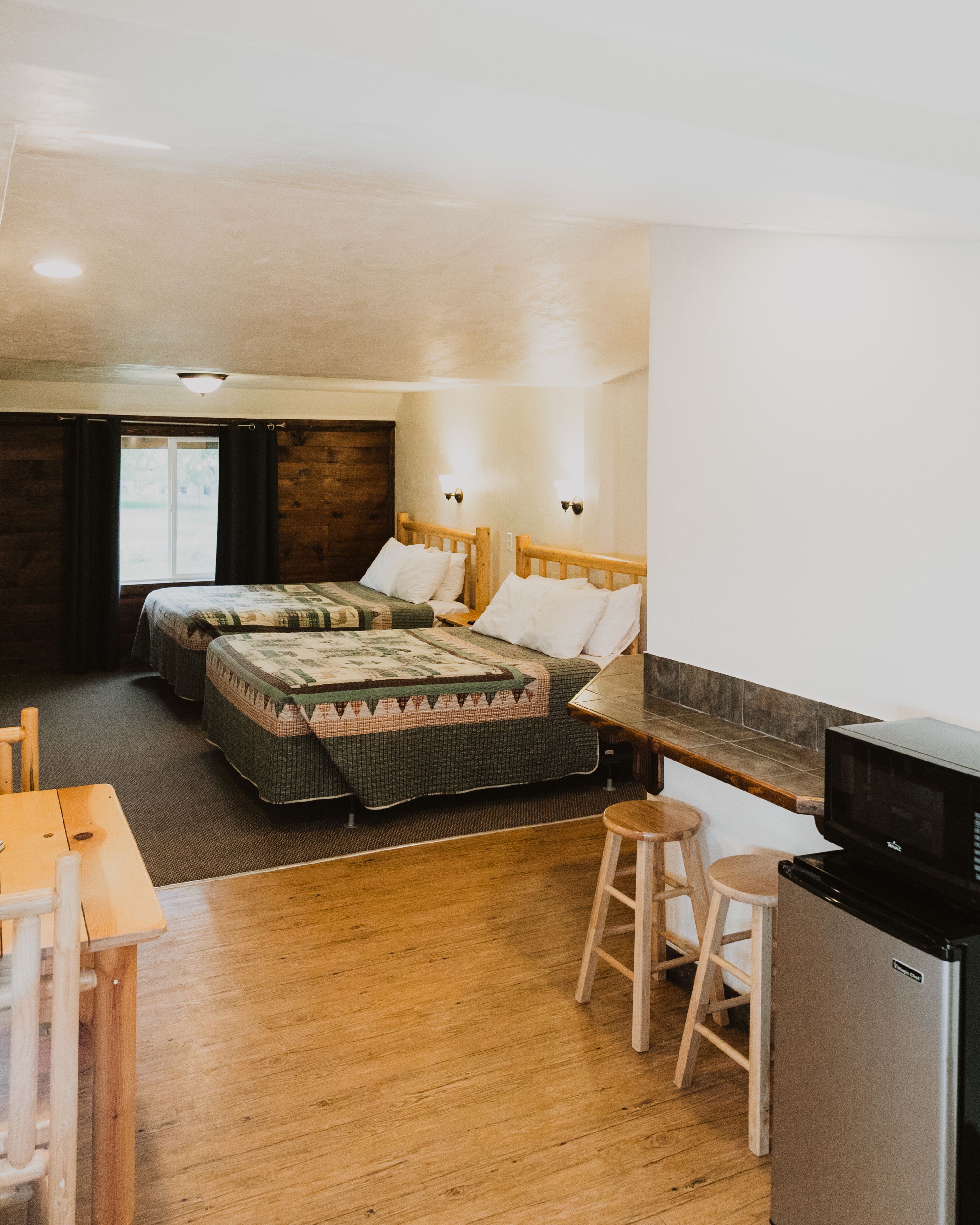 A compact hotel room with two beds, a kitchenette, a wooden dining table, and a TV.