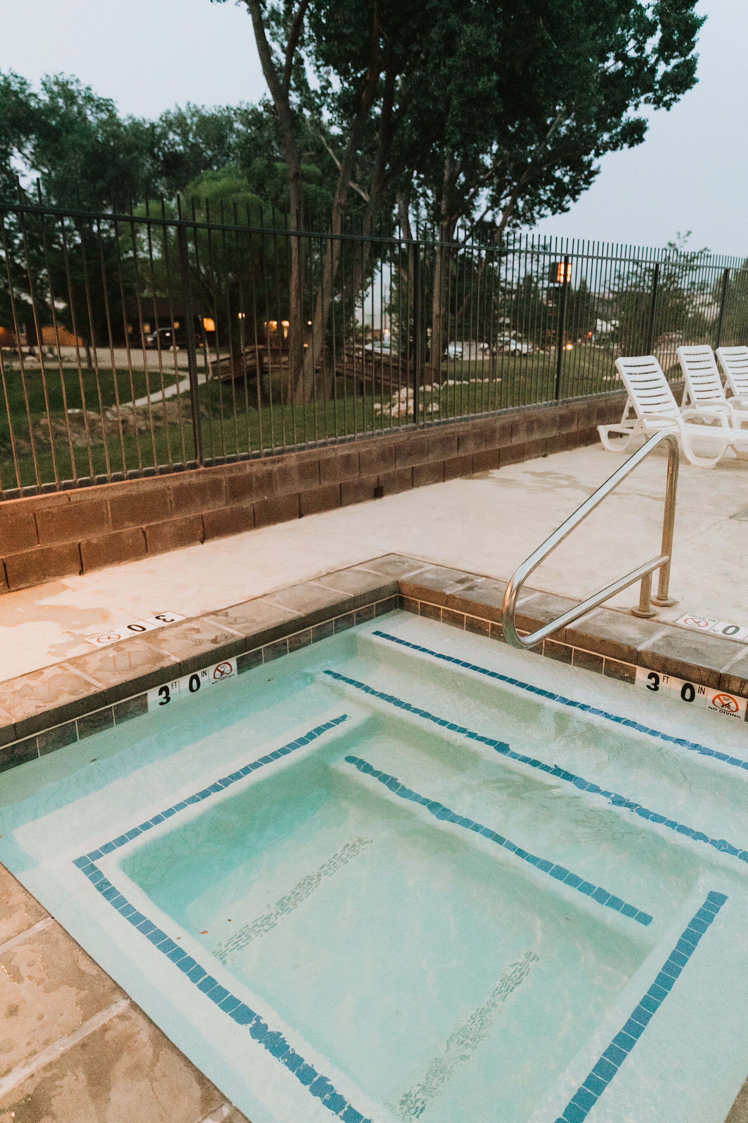 A swimming pool with a tiled edge, a metal ladder, and a fenced area with white lounge chairs.