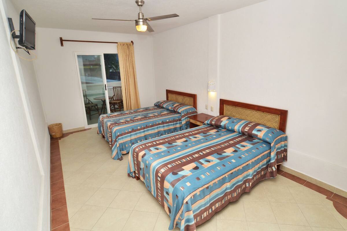 A hotel room with two beds, a ceiling fan, a TV mounted on the wall, and a sliding glass door leading to an outdoor area.