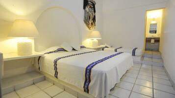Habitación de hotel con tres camas, ropa de cama blanca y colchas a rayas azules y amarillas. Cuenta con dos mesitas de noche con lámparas, un espejo grande y piso de baldosas.