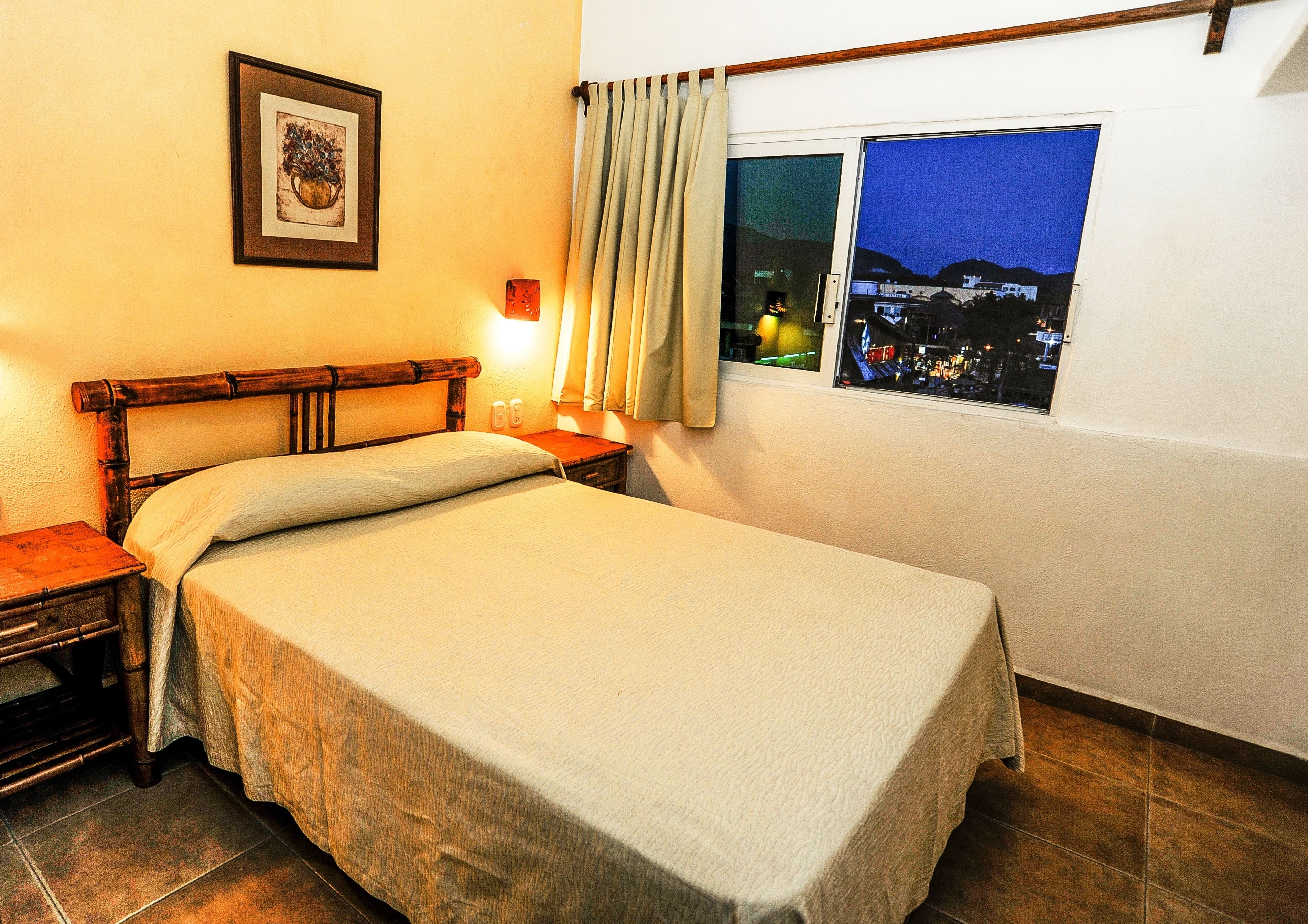 Una cama individual con cabecera de madera, una mesita de noche, un cuadro enmarcado en la pared, una ventana con vista a la ciudad de noche, y una cortina entreabierta.