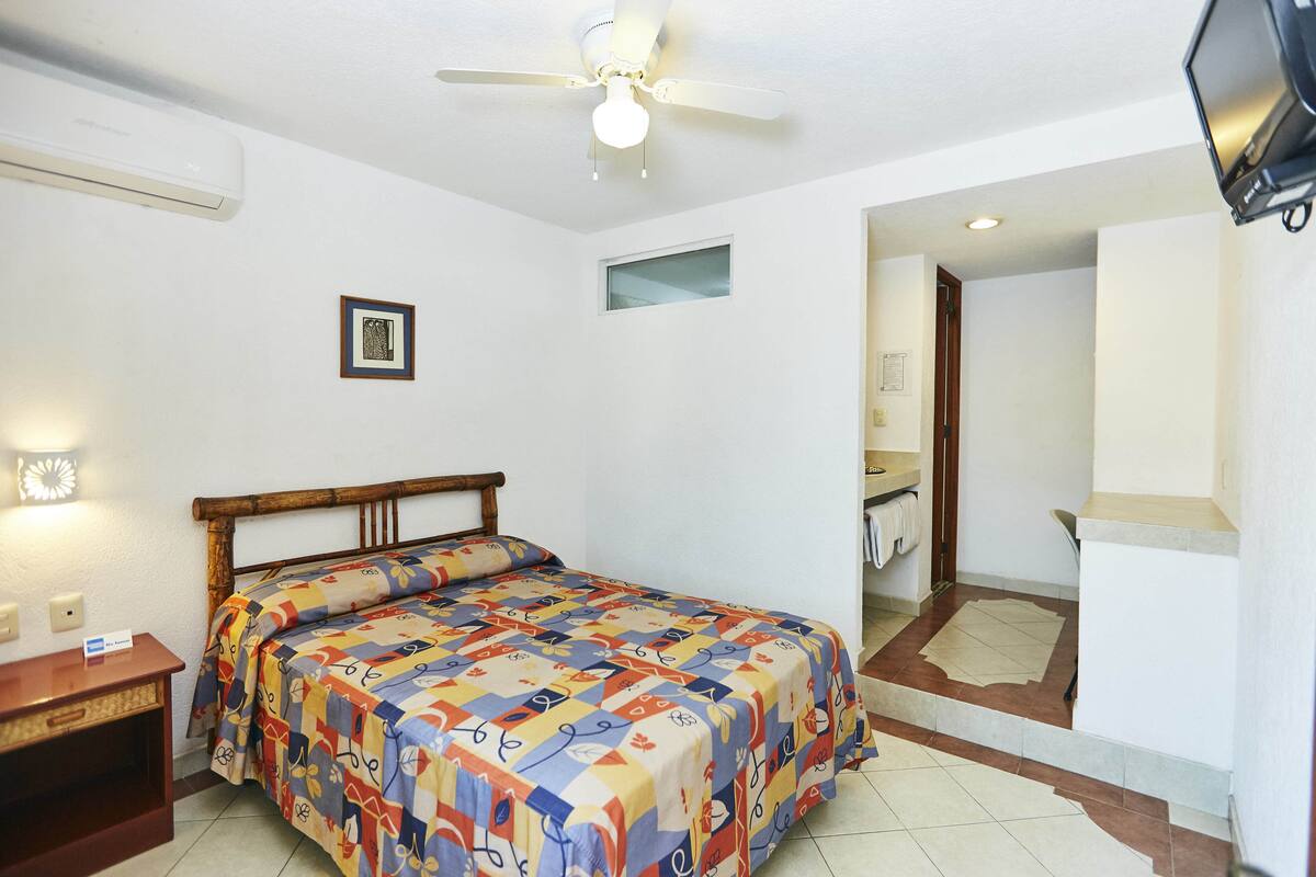 A bedroom with a bed, a ceiling fan, a small window, and a bathroom visible through an open door.
