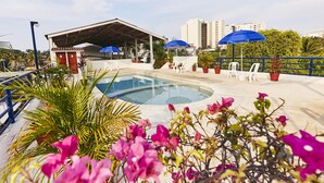 Una piscina con zona de estar cubierta, sillones reclinables blancos y sombrillas azules.
