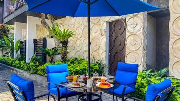 Un espace de restauration en plein air avec une table et quatre chaises bleues, un parasol et une variété d’aliments pour le petit-déjeuner.