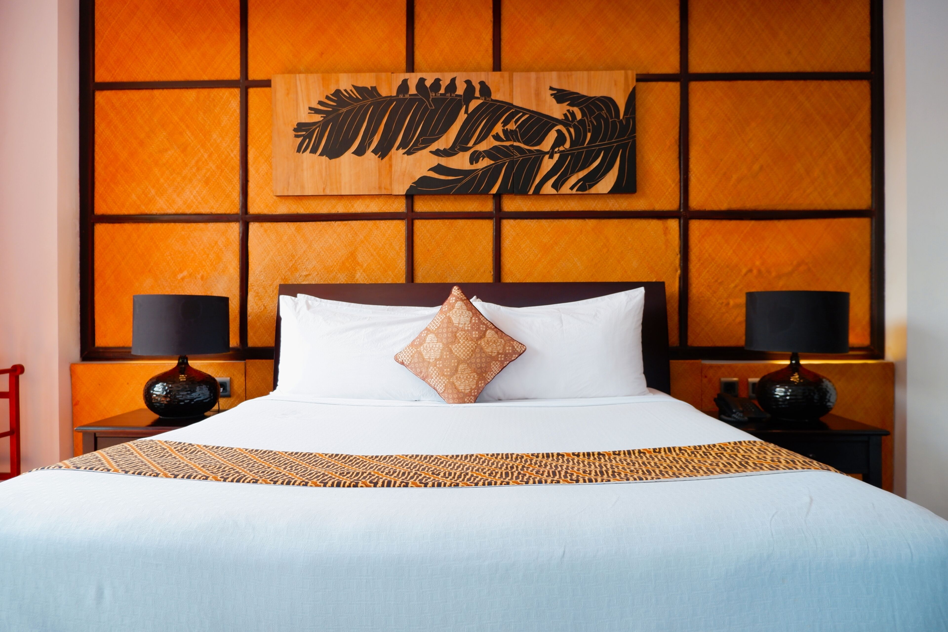 Habitación de hotel con una cama grande, dos mesitas de noche con lámparas, un panel decorativo en la pared y una silla roja.
