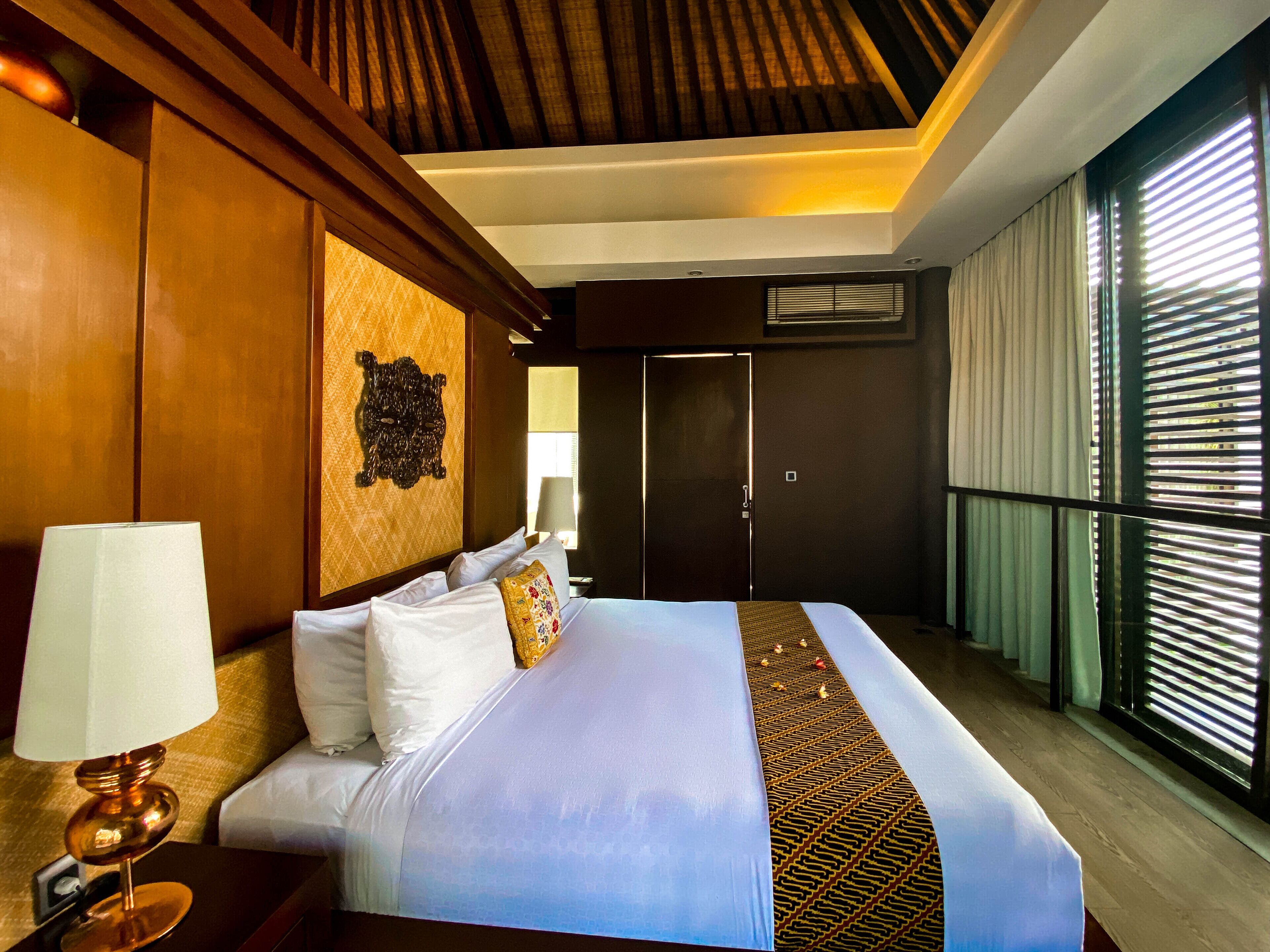 A hotel room with a large bed, a wooden headboard, a wall-mounted artwork, and a window with blinds.