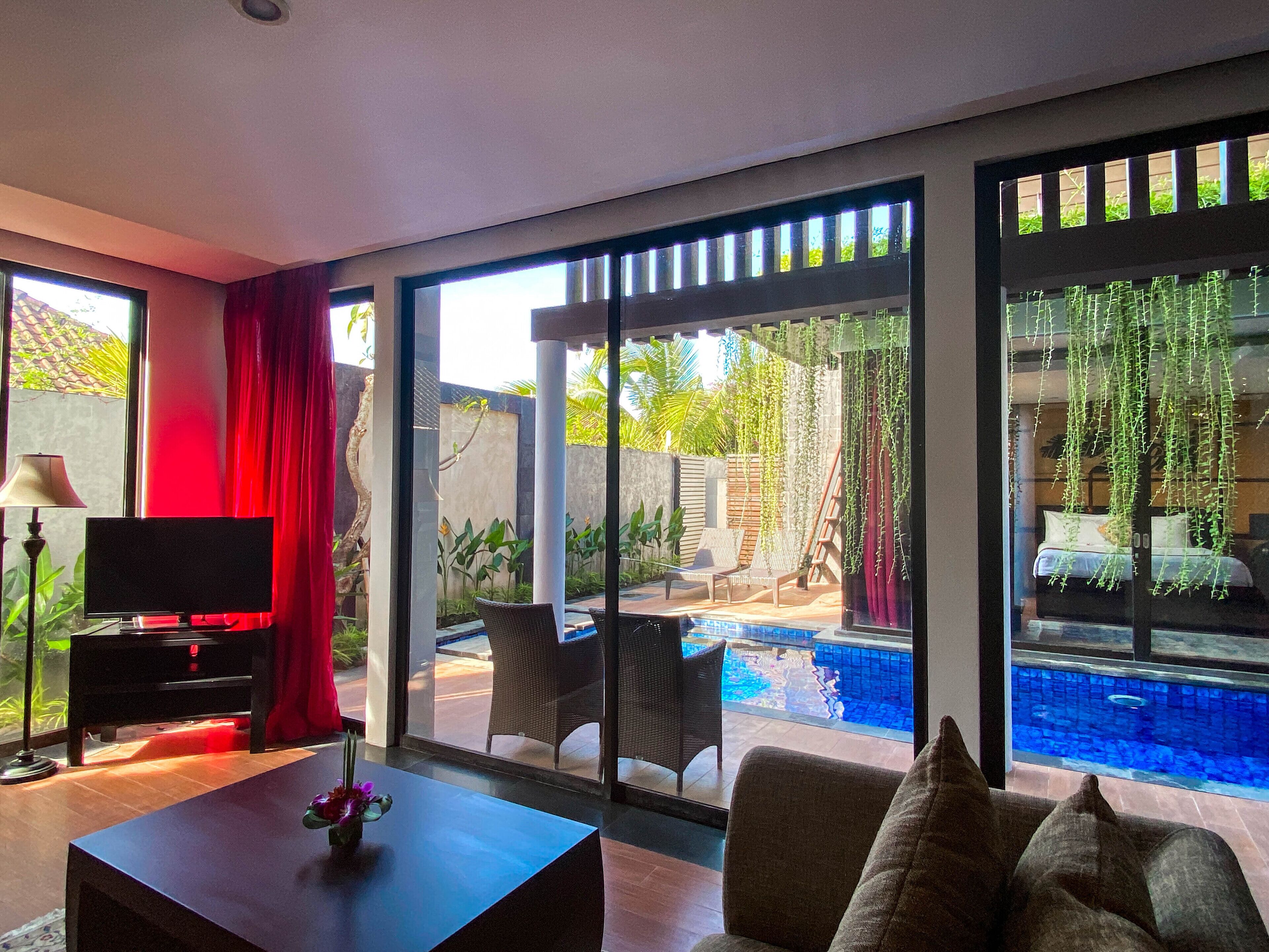 A modern living room with a TV, a coffee table with flowers, and a view of a swimming pool and outdoor seating area.