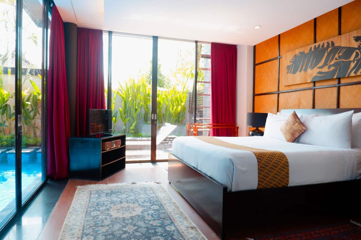 A modern hotel room with a large bed, a TV, a small table, and a view of a swimming pool and greenery.