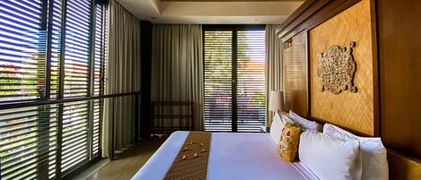 A large bed with a patterned bedspread, a wooden headboard with a decorative panel, and a view of greenery through floor-to-ceiling windows with blinds.